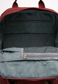 Bordowy plecak z pojemnym szarym wnętrzem, wyposażony w zamykaną na zamek kieszeń i siatkową przegrodę. Wytrzymały materiał, gładkie zamki błyskawiczne i wzmocnione szwy.