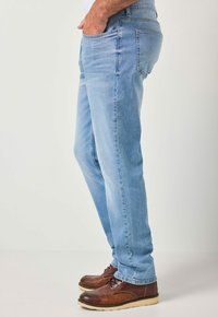 Mies, joka pukeutunut vaalean sinisiin farkkuihin ja ruskeisiin nahkakenkiin, seisoo sivuttain käsi taskussa yksiväristä taustaa vasten.