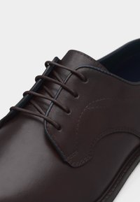 Κοντινό πλάνο σκούρου καφέ δερμάτινου παπουτσιού για επίσημες εμφανίσεις με στρογγυλά κορδόνια που περνούν από τέσσερις οπές και διακοσμητικό ράψιμο στο πλευρικό πάνελ.