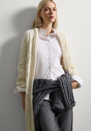 Cardigan in maglia spessa con motivo a treccia, indossato aperto su una camicia bianca con bottoni. Sciarpa grigia in maglia drappeggiata in vita, abbinata a jeans scuri.