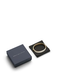 Zelta un sudraba bangles aproce, kas atpūšas uz melna auduma, blakus tumši zilai iepakojuma kastītei ar zelta tekstu, kurai ir tīrs, minimālistisks dizains.