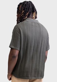 Olivgrünes Poloshirt mit kurzen Ärmeln, das ein strukturiertes Strickmuster mit horizontalen Streifen und kleinen Perforationen aufweist. Gerippter Kragen und Bündchen.