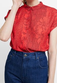 Červená blůza s hadím potiskem, zapínání na knoflíky, límec, krátké rukávy a lehká textura, kombinovaná s modrými džíny s vysokým pasem.