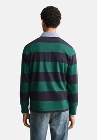 Ilgi piedurkņu polo krekls ar zaļu un tumši zilu svītru rakstu, ar apkakli un ribveduma aprocēm, kopā ar gaiši ziliem džinsiem.