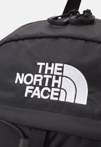 Rucsac negru din nailon, cu un logo alb brodat proeminent. Include accente hardware negre duale și detalii din țesătură texturată.