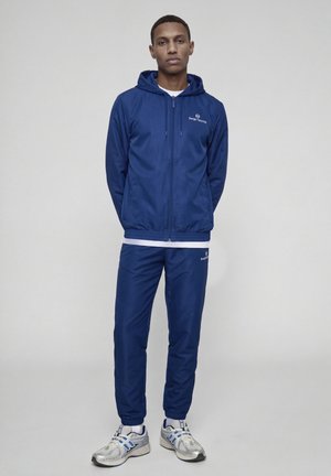 Un homme pose avec un sweat à capuche zippé bleu marine Sergio Tacchini et un pantalon de survêtement assorti, ainsi que des baskets blanches et bleues, sur un fond gris uni.