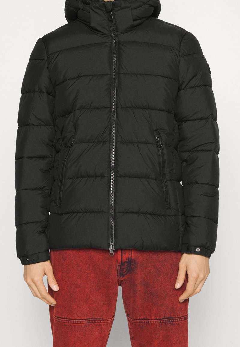 Veste matelassée noire avec capuche, fermeture éclair avant et poches latérales zippées, portée avec un pantalon en denim rouge délavé.