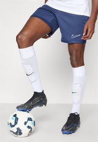 Σκαρπίνια ναυτικού τύπου με λευκό λογότυπο Nike, μαύρα ποδοσφαιρικά παπούτσια με μπλε λεπτομέρειες, λευκές κάλτσες μέχρι το γόνατο και μια υφασμάτινη λευκή ποδοσφαιρική μπάλα.