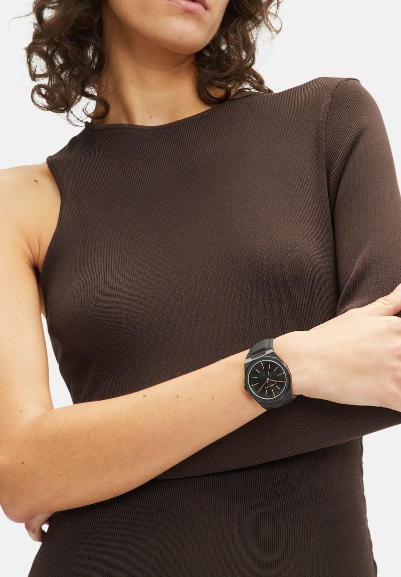 Bruin geribbelde top met een asymmetrische halslijn, gedragen met een zwart horloge met een ronde wijzerplaat en een matte band.