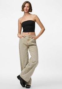 Giovane donna con capelli corti castani che indossa un top corto senza spalline nero, jeans beige a gamba larga e stivali neri alla caviglia, in piedi con le mani in tasca.
