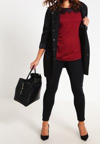 Kvinna som bär en svart kappa, bordeauxfärgad topp med spetsdetaljer, svarta leggings, svarta klackar och bär en stor strukturerad svart handväska.