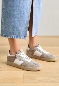 Pieds portant des baskets en suède marron clair avec des rayures blanches et des lacets blancs, associés à un pantalon en denim bleu clair avec une fente arrière.