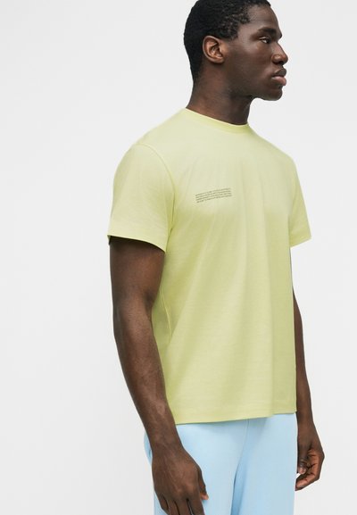 Άνδρας που φορεί κοντομάνικη μπλούζα ανοιχτού κίτρινου χρώματος με μικρό τυπωμένο κείμενο στο στήθος, σε συνδυασμό με ανοιχτό μπλε παντελόνι, στέκεται πλαγίως.