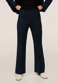 Navy blue trousers with a relaxed fit, featuring sharp creases, a high waistband, and side pockets. Worn with a black top and light shoes.