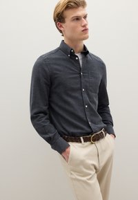 Camisa de color gris oscuro abotonada, con un bolsillo en el pecho, combinada con pantalones de color claro y un cinturón trenzado marrón con hebilla de bronce.