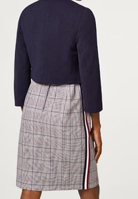 Giacca corta blu navy con maniche a tre quarti abbinata a una gonna grigia a quadri con dettaglio laterale a righe contrastanti.