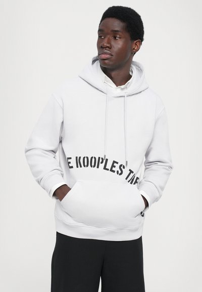 Junger Mann in weißem Hoodie mit schwarzem Text und schwarzen Hosen, Hände in der vorderen Tasche, schaut nach links vor einem schlichten Hintergrund.