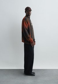 Gebreide cardigan in een oranje-bruine gradient, met knopen en lange mouwen, gecombineerd met een donker T-shirt en zwarte broek. Zwarte schoenen gedragen.