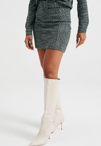 Falda mini a cuadros verde y blanco con un diseño ajustado, combinada con botas de cuero blanco de punta afilada hasta la rodilla.