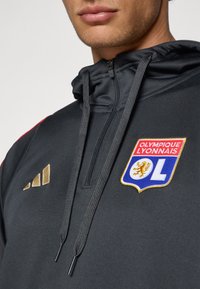 Sort hættetrøje med snore; har gyldne Adidas-striber og broderet Olympique Lyonnais-logo i blå, rød og guld.