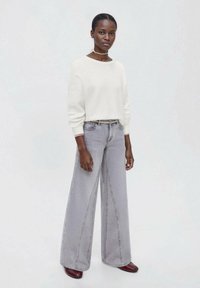 Bela teksturirana pulover s prostim krojem, kombiniran z visokimi sivo-robnimi hlačami širokega kroja. Model nosi temno rdeče čevlje in stoji na svetli podlagi.
