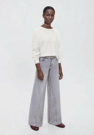 Pull blanc texturé à coupe décontractée, associé à un jean gris taille haute à jambe large. Le mannequin porte des chaussures rouge foncé, se tenant devant un fond clair.