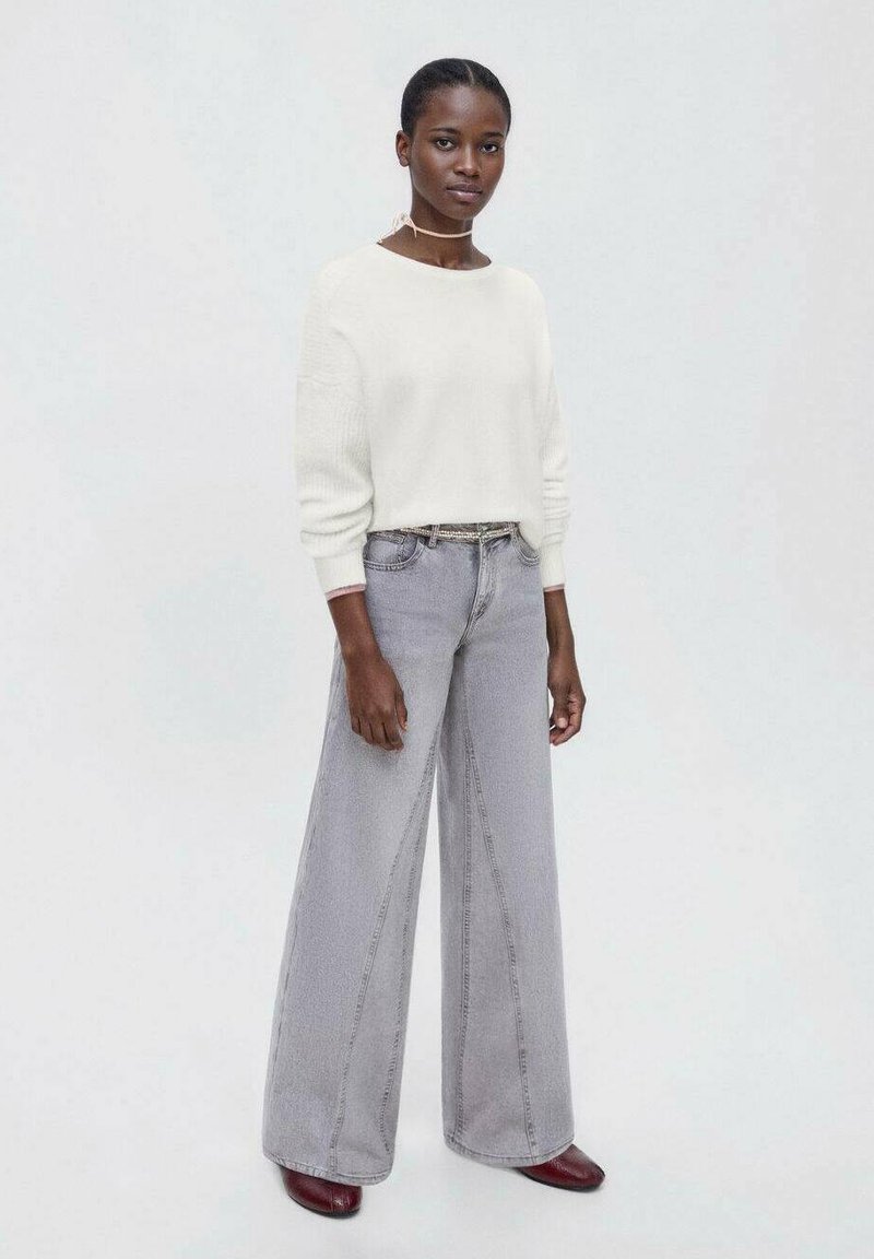 Bela teksturirana pulover s prostim krojem, kombiniran z visokimi sivo-robnimi hlačami širokega kroja. Model nosi temno rdeče čevlje in stoji na svetli podlagi.