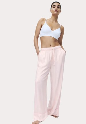 Donna con indosso un bralette bianco e pantaloni larghi rosa pallido con coulisse, in piedi scalza con le mani nelle tasche contro uno sfondo neutro.