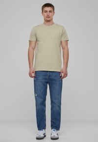 Camiseta de algodón beige combinada con jeans azules desgastados. El modelo lleva deportivas grises y blancas con acentos azules. Diseño simple y casual.