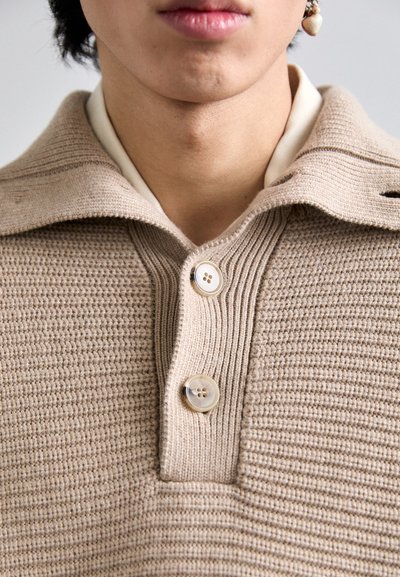 Beige knitted polo shirt featuring a textured weave, three buttons, and a fold-over collar, with a layered cream shirt visible beneath.
