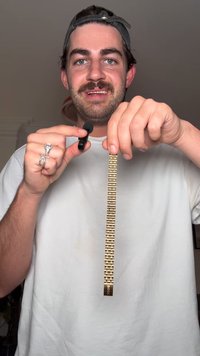 Kultainen metallinen kellonranneke suorakulmaisilla linkeillä ja lukolla, jossa on kaiverrettu "BOSS"; henkilö pitää sitä, ja hänellä on valkoinen paita päällä.