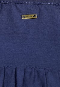 Un tissu bleu marine à surface texturée présente une petite étiquette dorée "RAW" fixée, accentuant le détail de couture froncé en dessous.
