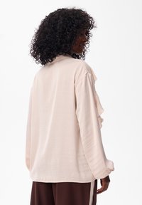 Kobieta z kr�ęconymi włosami, ubrana w luźną, bladoróżową bluzkę z długim rękawem i ciemno-brązowe spodnie, stoi bokiem na jednolitym tle.