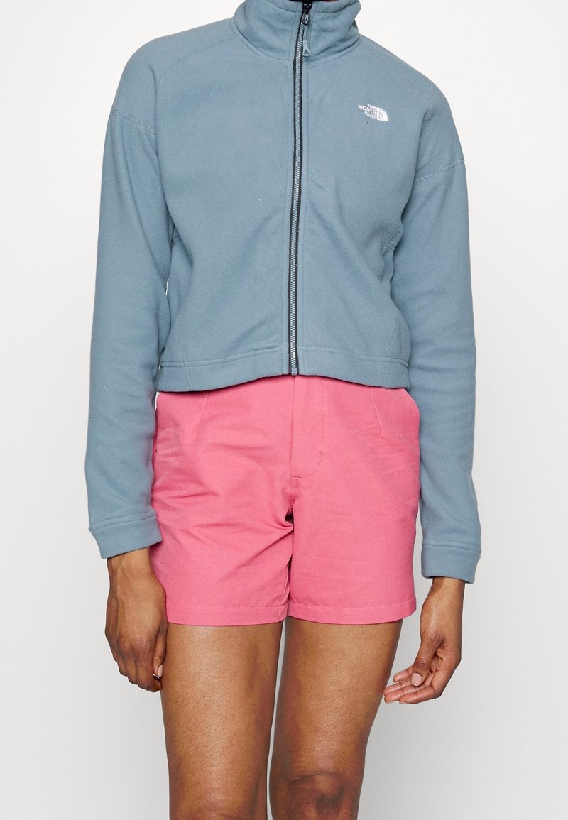 Chaqueta de forro polar cortada en azul claro con cuello alto y cremallera. Combinada con pantalones cortos rosas. Ambas prendas tienen un ajuste holgado.