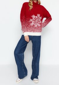 Punainen neulepaita, jossa on valkoinen lumihiutale-kuviointi, gradienttikuosi, korkea kaulus ja taitetut hihat, yhdistettynä leveälahkeisiin sinisiin farkkuihin ja valkoisiin lenkkareihin.