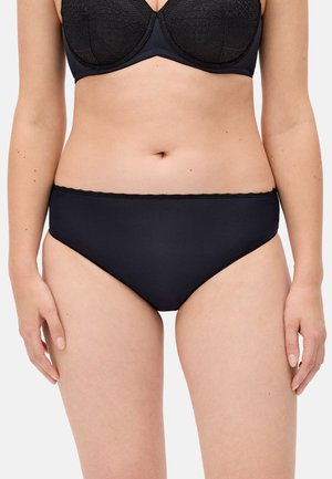 Mujer con sujetador de encaje negro y braguitas negras a juego, de pie contra un fondo liso, vista frontal recortada a la altura de los hombros y los muslos.