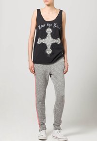 Schwarzes Tanktop mit weißem Grafikdruck eines Kreuzes, kombiniert mit grauen Sweatpants mit rosa Seitenstreifen und weißen Turnschuhen.