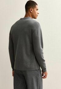 Ung man klädd i en grå långärmad tröja och rutiga byxor, stående på sidan mot en ljus enfärgad vägg.