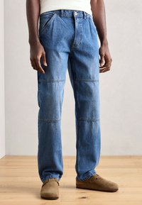 Lyseblå denim jeans med rett ben-design, med frontlommer og tonalt søm. Båret med brune slip-on sko på et tregulv.