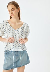 Witte blouse met blauwe driehoekpatronen, pofmouwen en trekkoordhalslijn. Gecombineerd met een lichte denim minirok.