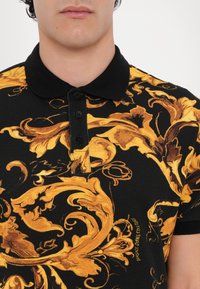 Camisa polo preta com padrão floral ornamentado em amarelo e castanho, apresentando um colarinho preto e uma pala com quatro botões. O material parece ter textura.