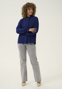 Mörkblå blus med volangkrage och knäppning framtill, i kombination med ljusgrå jeans. Blusen har ballongärmar och en avslappnad passform.