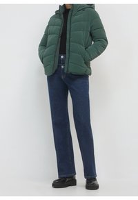 Giacca a vento verde con cappuccio, caratterizzata da una texture trapuntata e una chiusura frontale con zip, abbinata a jeans in denim scuri a vita alta.