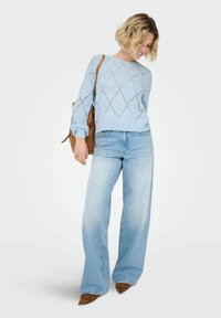 Pull en tricot bleu clair avec motif en losanges, associé à un jean large en denim clair. Le mannequin tient un sac à main marron. Chaussures pointues marron.