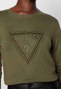 Maglione a maglia verde oliva con un grande logo a triangolo caratterizzato da cuciture intricate e accenti metallici. Collo rotondo e polsini a coste.