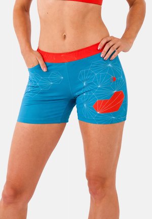 Femme portant un short de sport bleu avec des accents rouges et des motifs géométriques en lignes blanches, posant avec les mains sur les hanches contre un fond blanc.