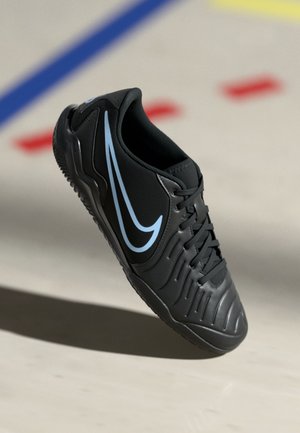 Sort atletiksko med lyseblå Nike-swoosh på siden, snørebånd bundet, fotograferet i luften over en lys gulv med farvede linjer.