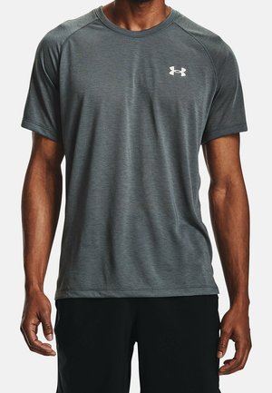 Mann trägt ein graues Under Armour Sportshirt mit kurzen Ärmeln und schwarze Shorts, steht mit locker an den Seiten hängenden Armen.