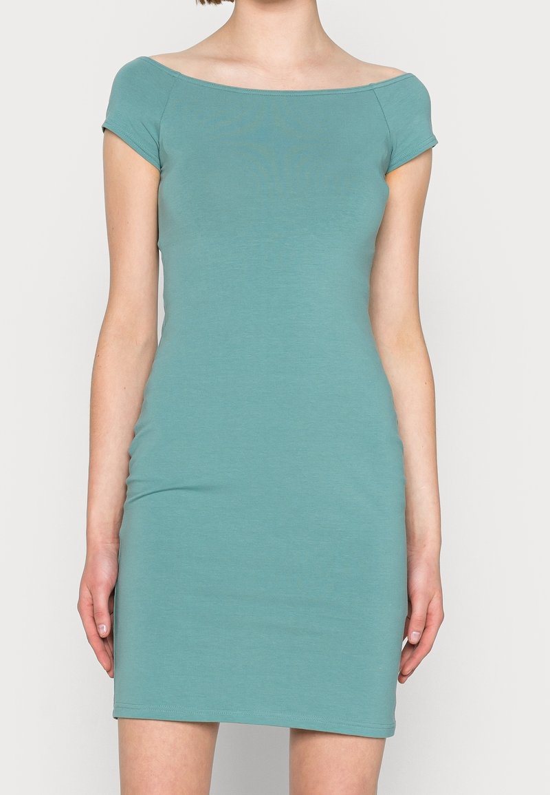 Robe courte ajustée à manches courtes de couleur sarcelle avec un large décolleté bateau, portée par une personne debout devant un fond uni.
