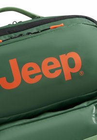 Mochila verde con gran logo naranja de "Jeep", tela texturizada, acentos negros y tirador de cremallera naranja, con múltiples compartimentos.
